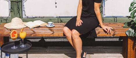 Una persona sentada en un banco de madera con una mesa y bebidas frente a ella, con vistas a un paisaje urbano que incluye edificios y montañas al fondo.