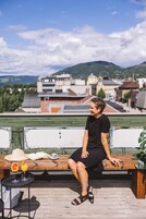Uma pessoa sentada num banco de madeira, com uma mesa e bebidas à frente, com vista para uma paisagem urbana com edifícios e montanhas ao fundo.