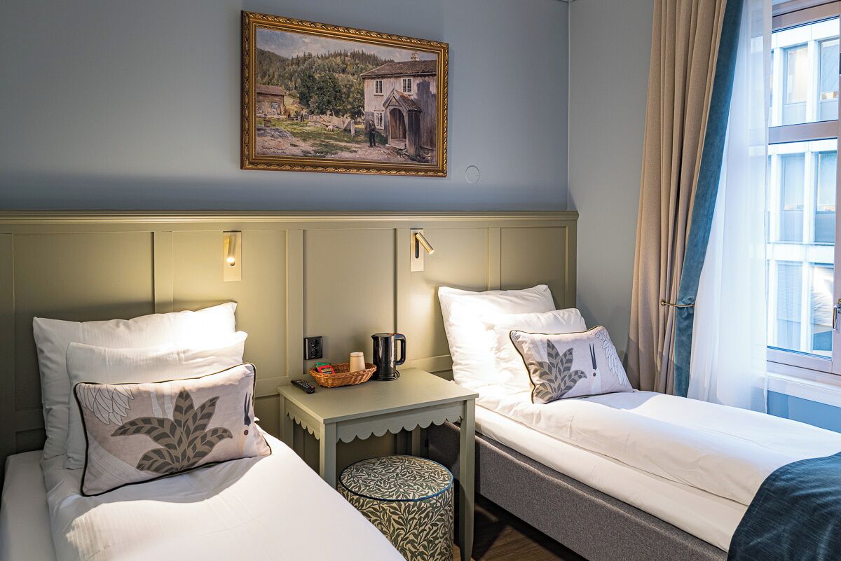 A hotel room with two single beds, a small table with a basket and a kettle, a painting on the wall, and a window with curtains.