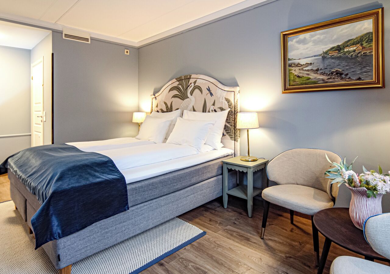 Un dormitorio ordenado con cama, lámparas de noche, una silla y una mesita con un jarrón de flores.