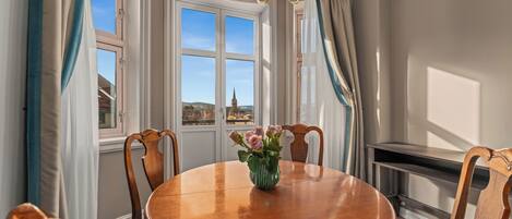 A dining area with a round wooden table, chairs, a vase with flowers, and a view of a building through large windows.