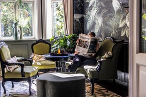 Un hombre leyendo un periódico en una habitación con un ventanal grande, una mesa redonda con una planta y un cuadro en la pared.