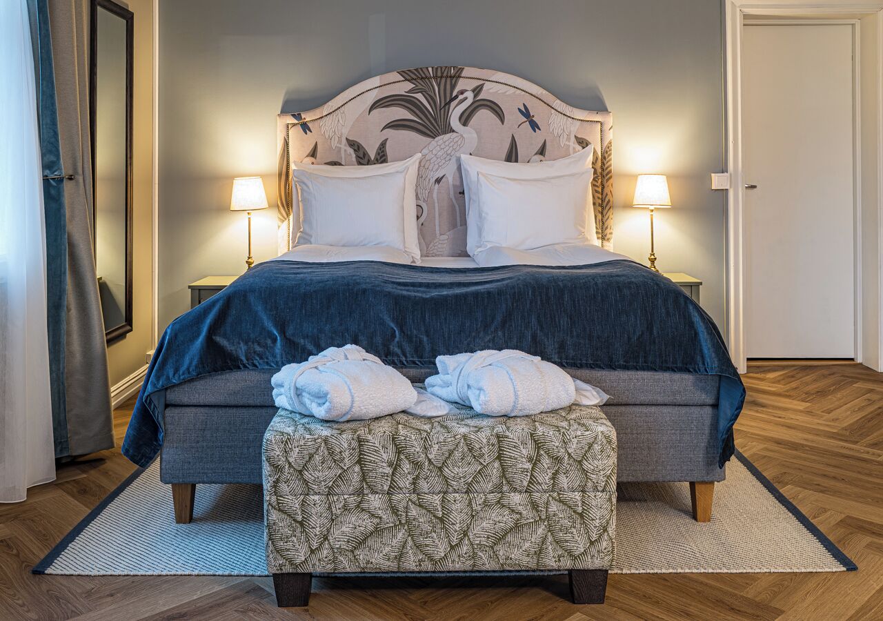 A bed with a headboard, two bedside lamps, a bench with towels, and a patterned rug.