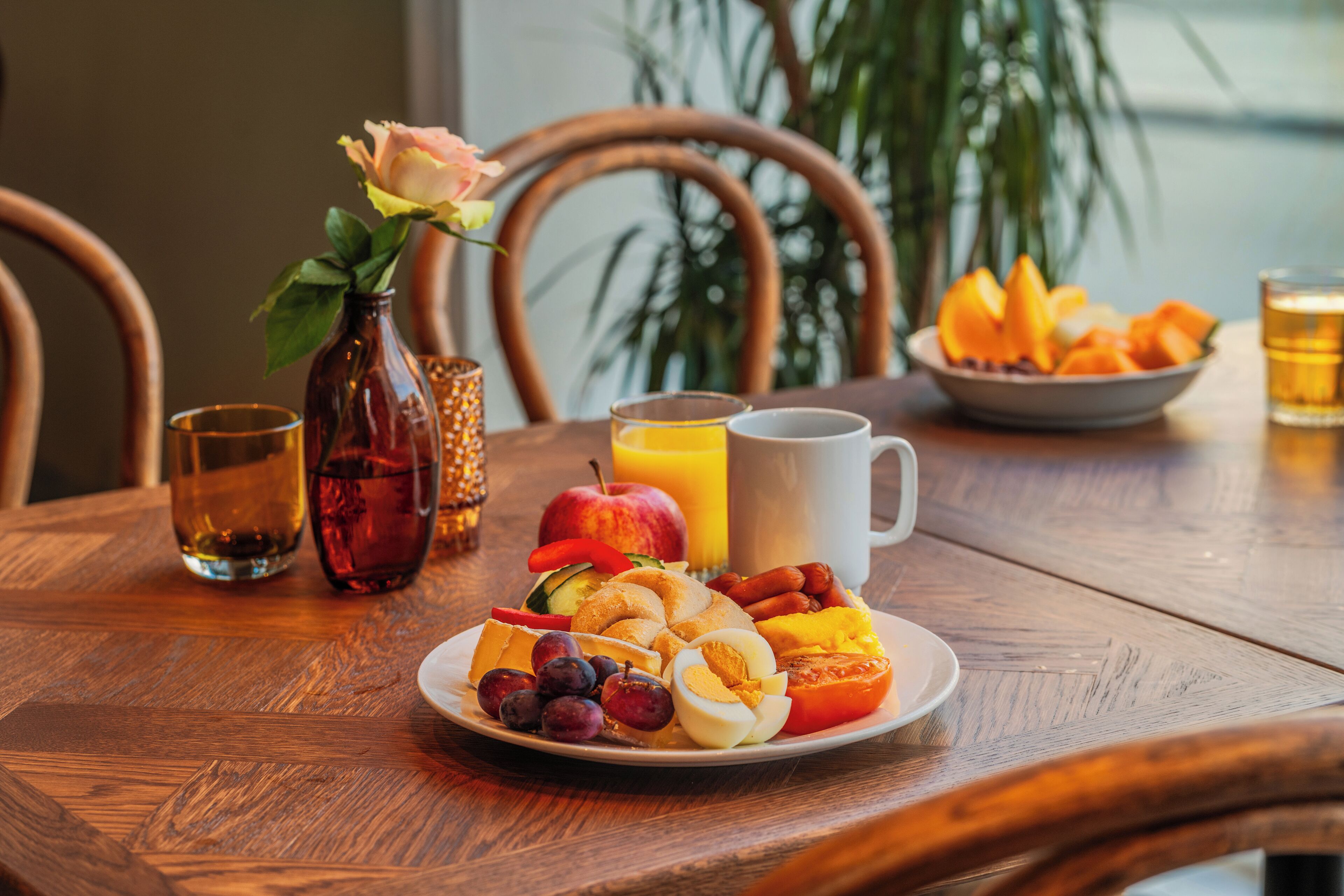 A breakfast setting with a plate of fruits, eggs, and sausages, a bottle of juice, and a glass of beer on a wooden table.