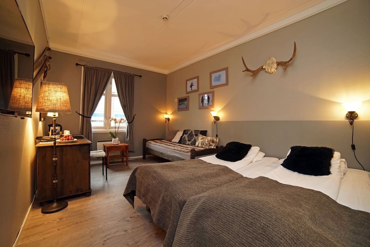 A bedroom with two beds, a desk with a lamp, a chair, and a window with curtains.
