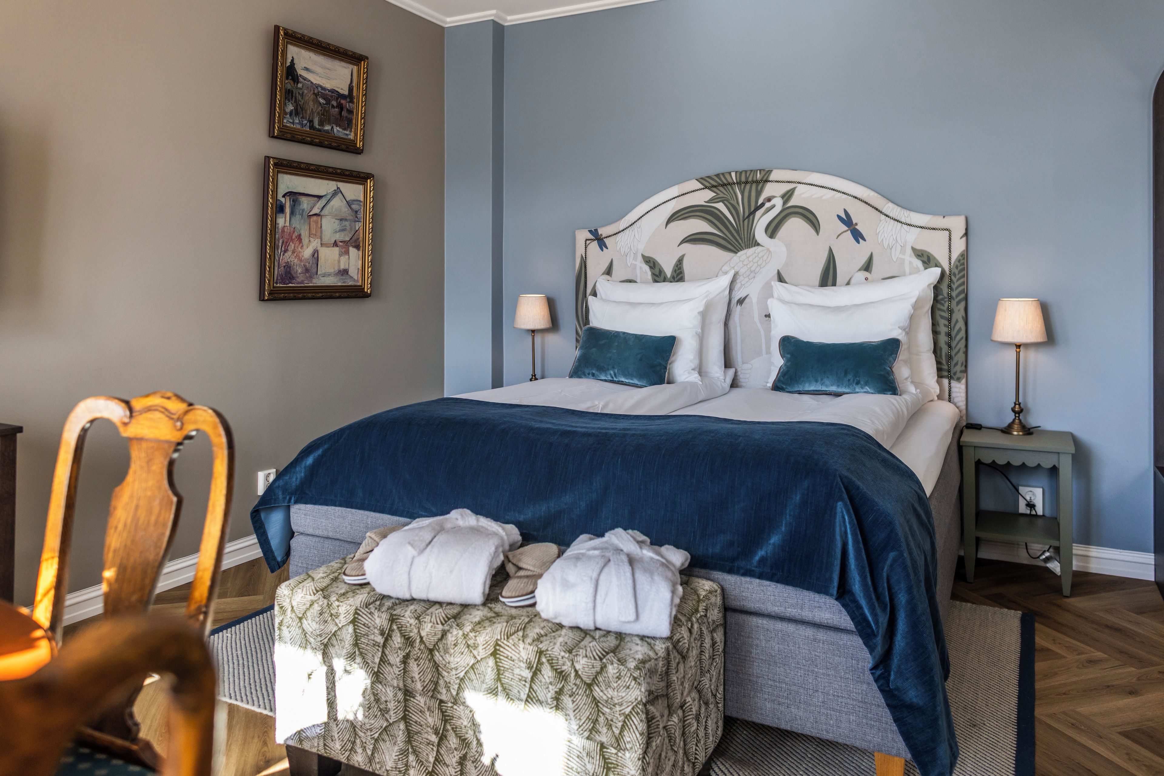 A neatly made bed with a blue headboard, two bedside lamps, and a set of towels on a chair.