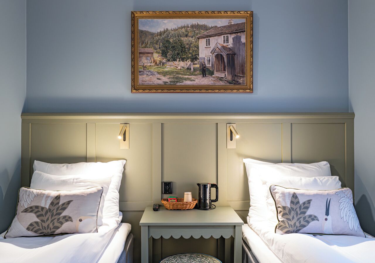 Een kamer met twee bedden, een klein tafeltje met een waterkoker, een mand en een lijst met een schilderij aan de muur.