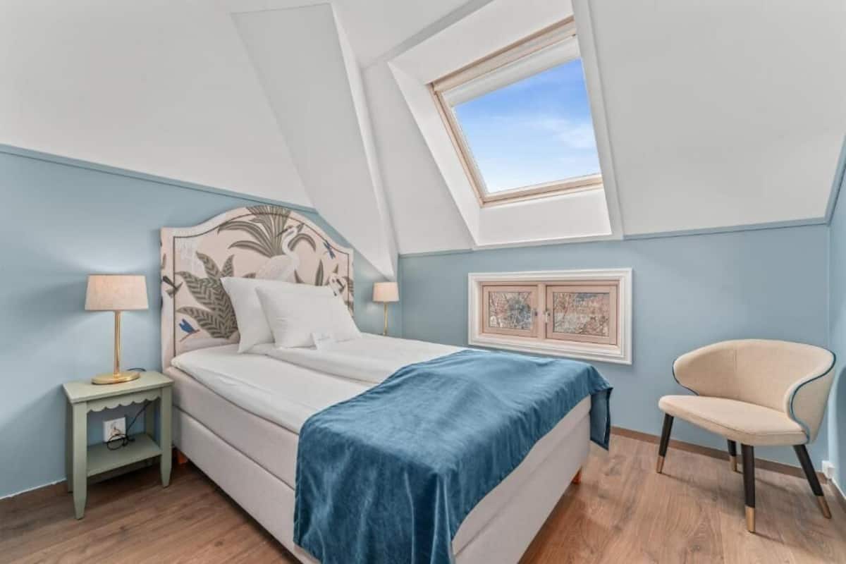 A bedroom with a bed, a nightstand, a chair, a window, and a picture frame on the wall.