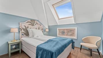 Un dormitorio con cama, mesita de noche, silla, ventana y un cuadro en la pared.