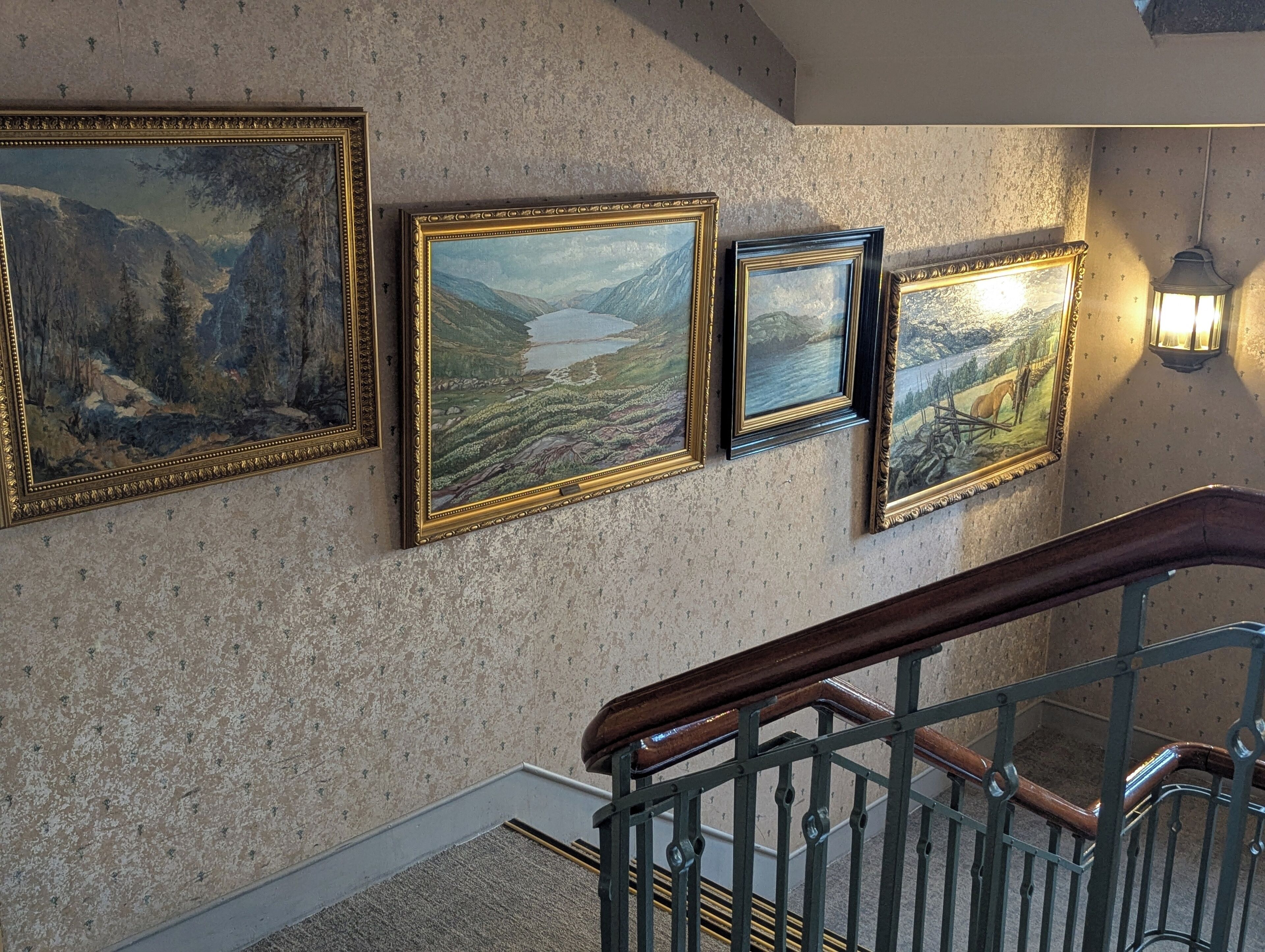 A staircase with a wooden handrail and metal balusters, leading to a landing with three framed landscape paintings on a textured wall.
