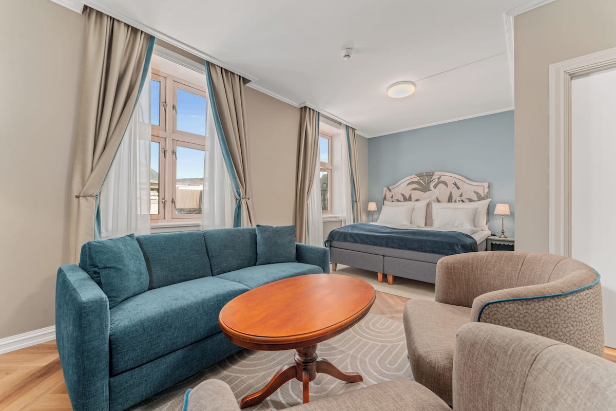 A modern hotel room with a blue sofa, a wooden table, a grey bed, and a patterned armchair.