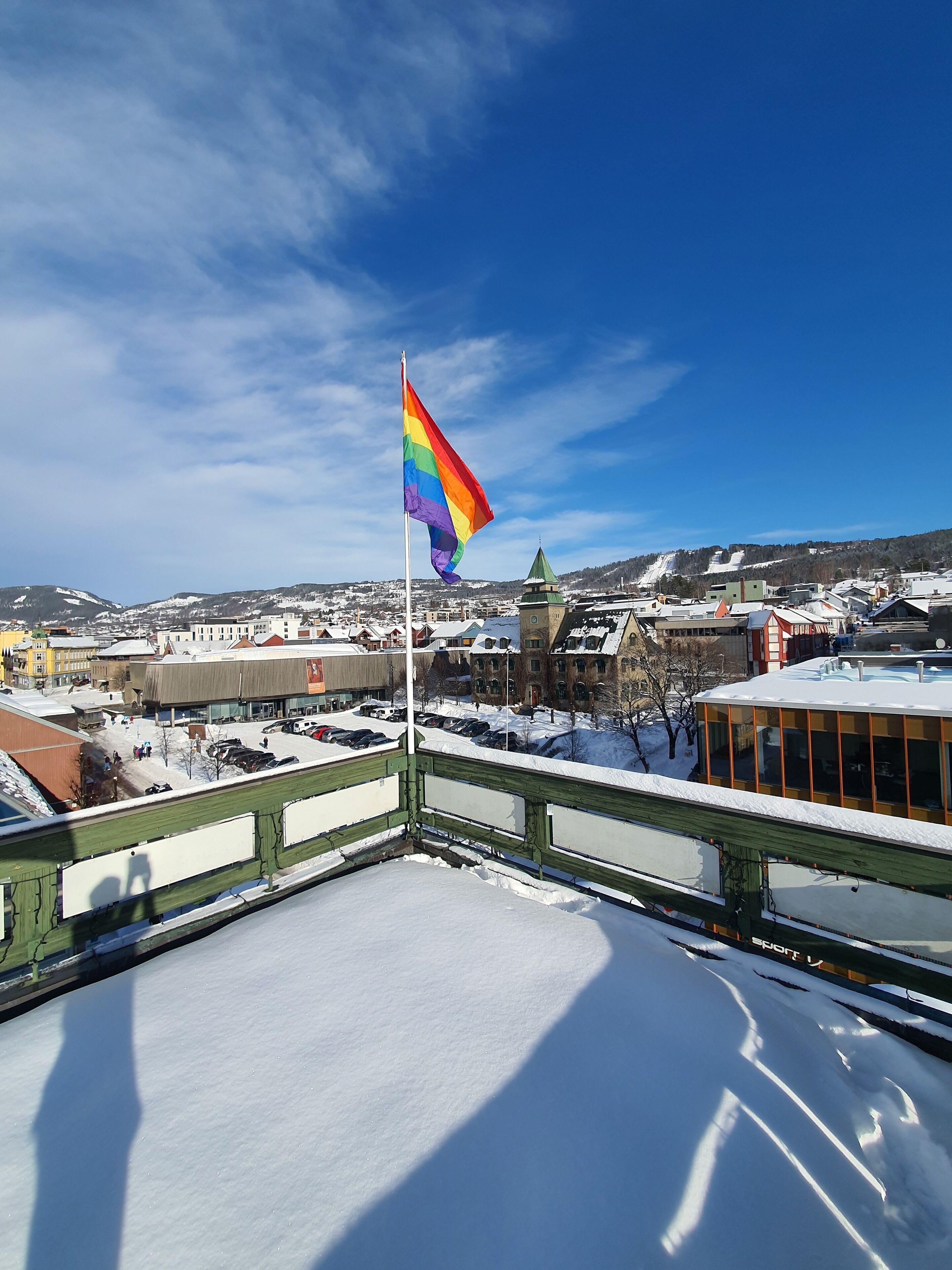 Paisatge nevós amb una bandera arcoiris a un penya-segat, edificis i un cel blau clar.