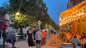 Una calle empedrada bordeada de árboles y edificios, con gente caminando y cenando al aire libre.
