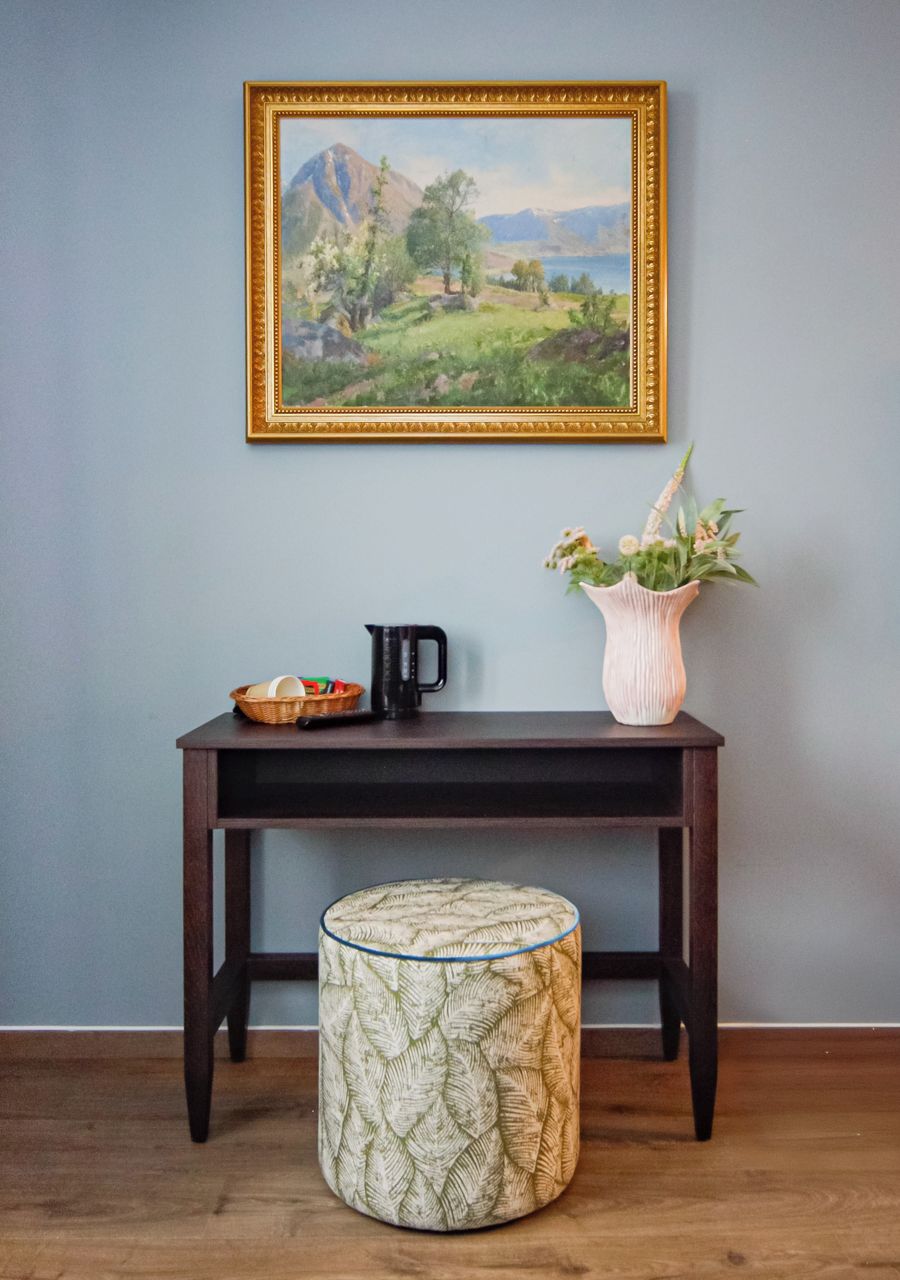 Una mesa de madera oscura con un cuadro, un florero con flores, una tetera y un reposapiés estampado frente a una pared azul claro.