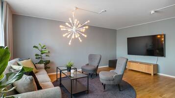 Un salon moderne avec un canapé gris, deux fauteuils, une table basse, un téléviseur et un luminaire suspendu au plafond.