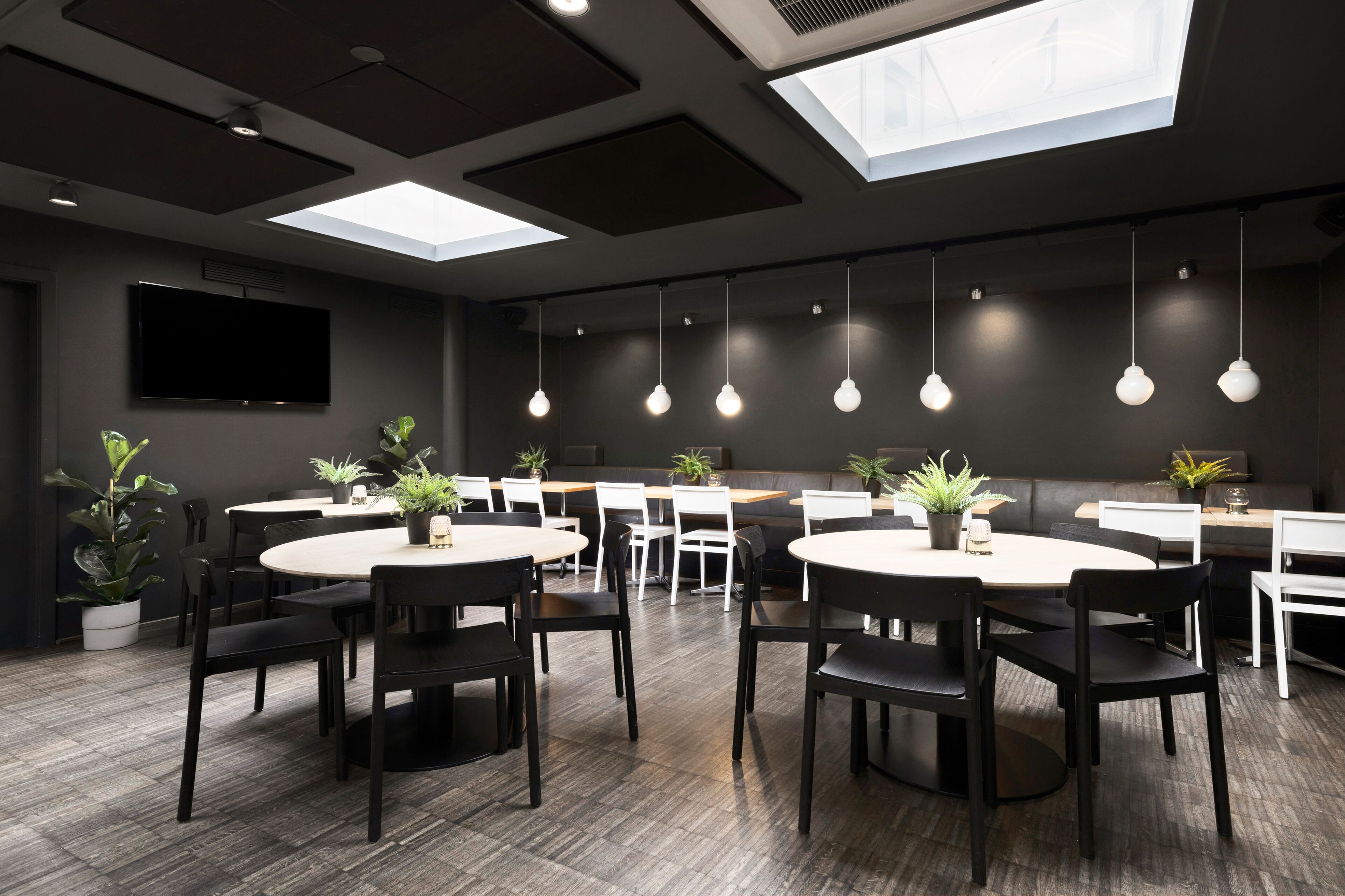 Une salle de conférence moderne, dotée de tables rondes, de chaises noires, de plantes en pot et d’un téléviseur à écran plat.