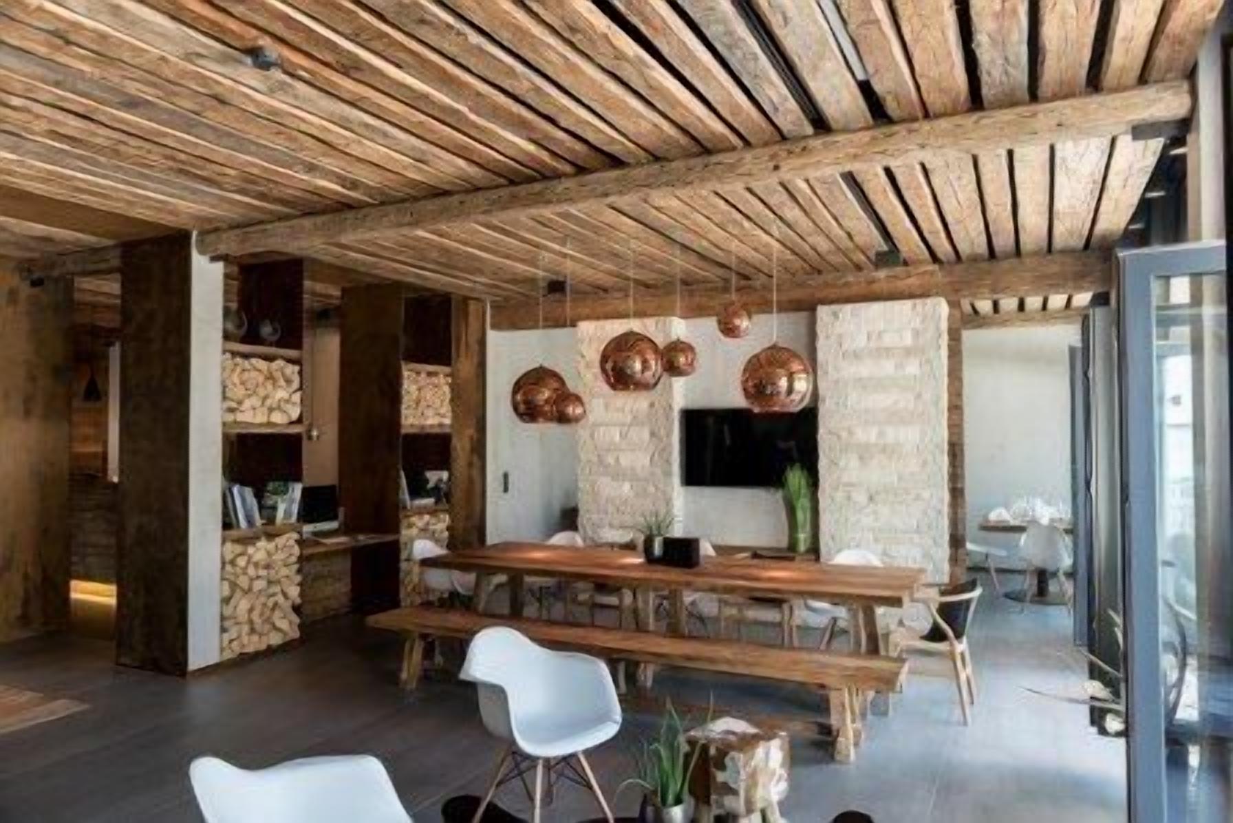 A rustic interior with exposed wooden beams, a long wooden table, white chairs, and copper pendant lights.