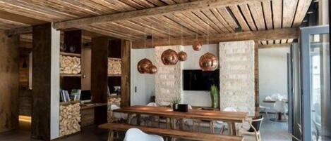 A rustic interior with exposed wooden beams, a long wooden table, white chairs, and copper pendant lights.