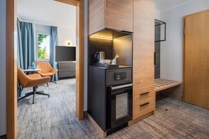Quarto de hotel compacto com cozinha compacta, área de estar com duas cadeiras, televisão e janela com cortinas.