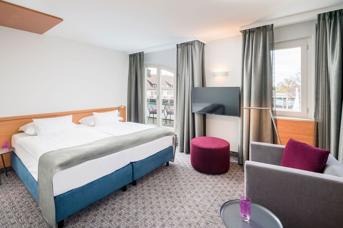 A hotel room with a bed, a TV, a seating area, and a balcony with a view.