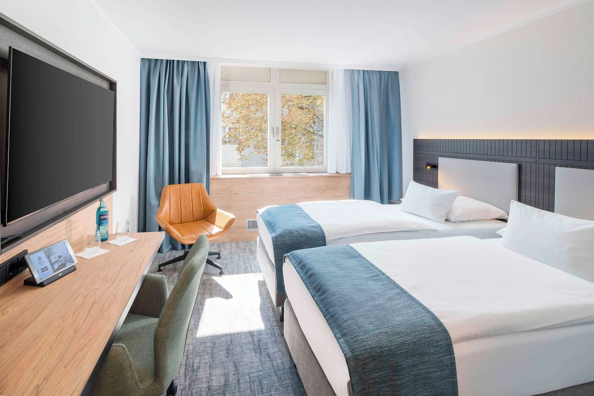 A hotel room with two beds, a desk with a TV, a chair, and a large window with curtains.