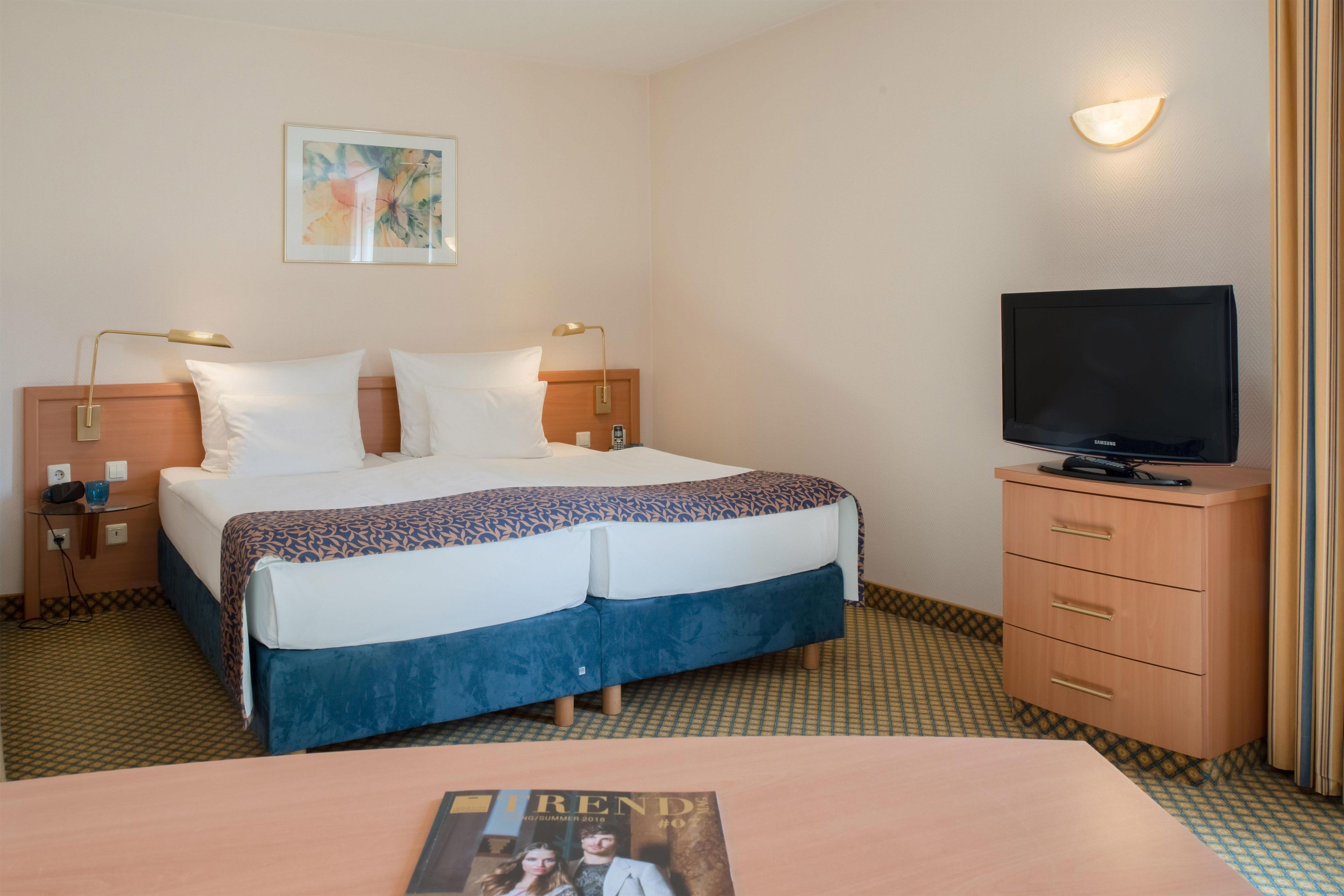 Een hotelkamer met een bed, nachtlampjes, een televisie, een bureau en een tijdschrift op de tafel.