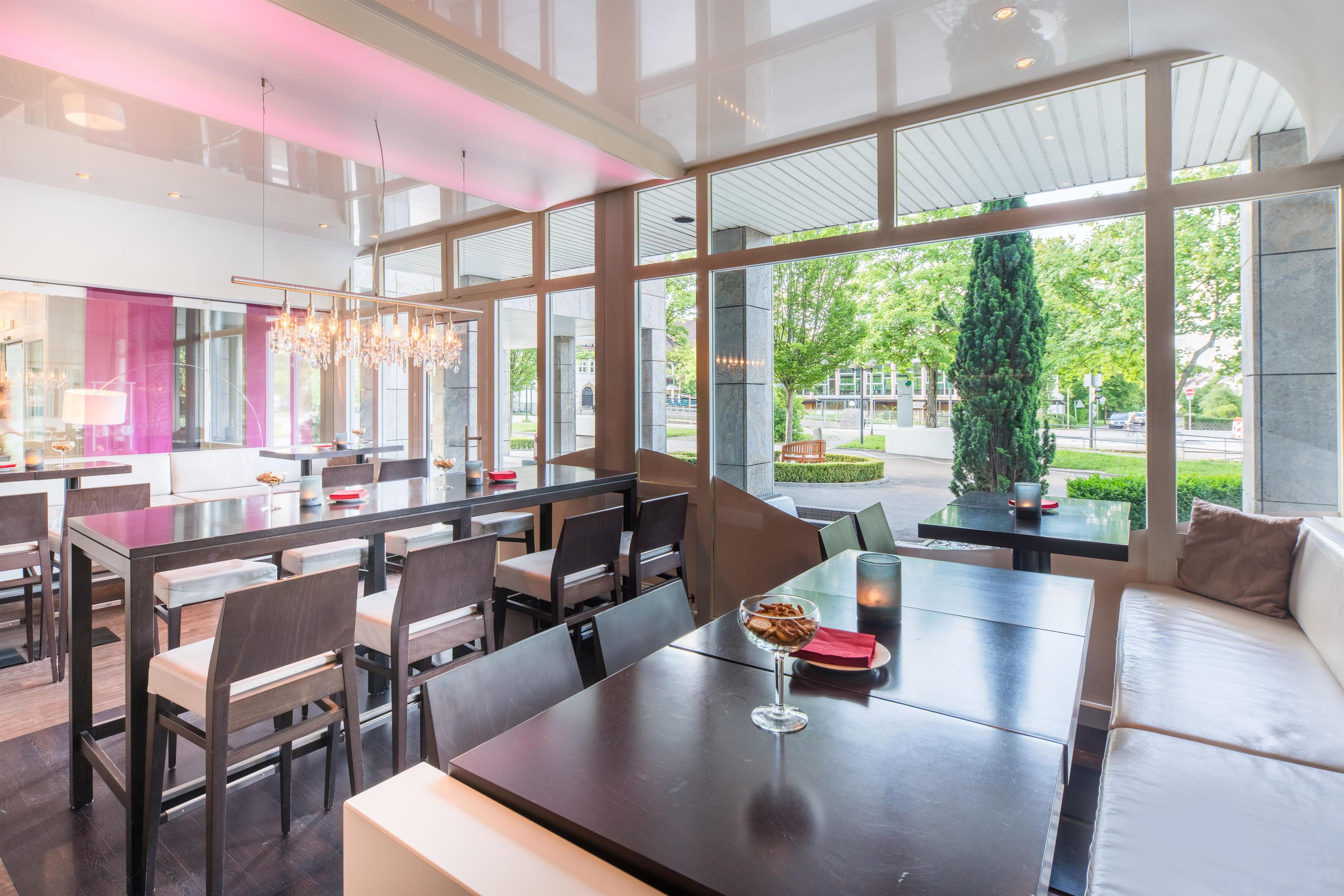 Ein modernes Restaurant mit hohen Decken, großen Fenstern und einer Kristallleuchter. Tische sind für das Essen angerichtet.