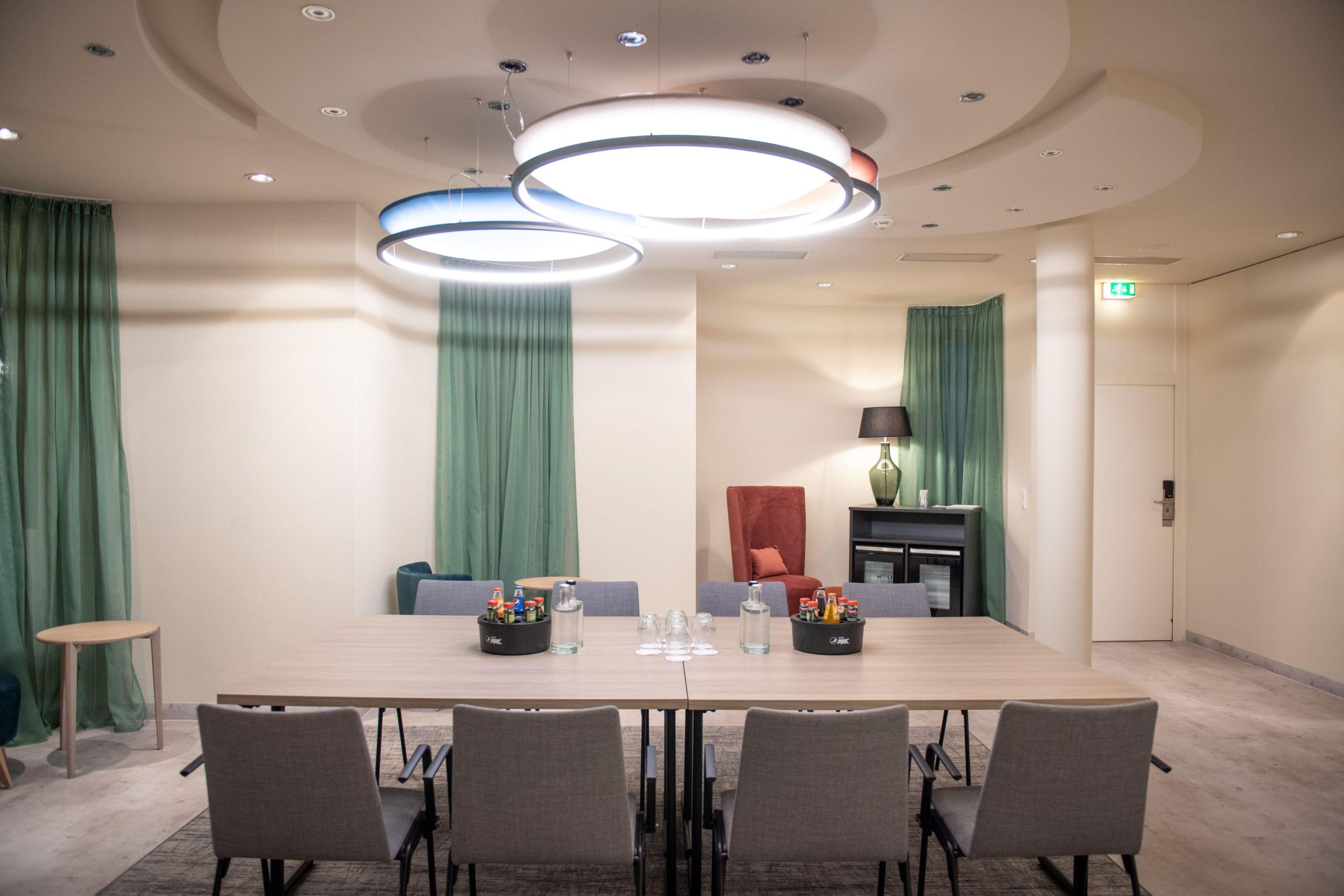 Ein Konferenzraum mit einem langen Tisch, sechs Stühlen, einem kleinen Beistelltisch mit Flaschen und einer Lampe auf einem Seitentisch.