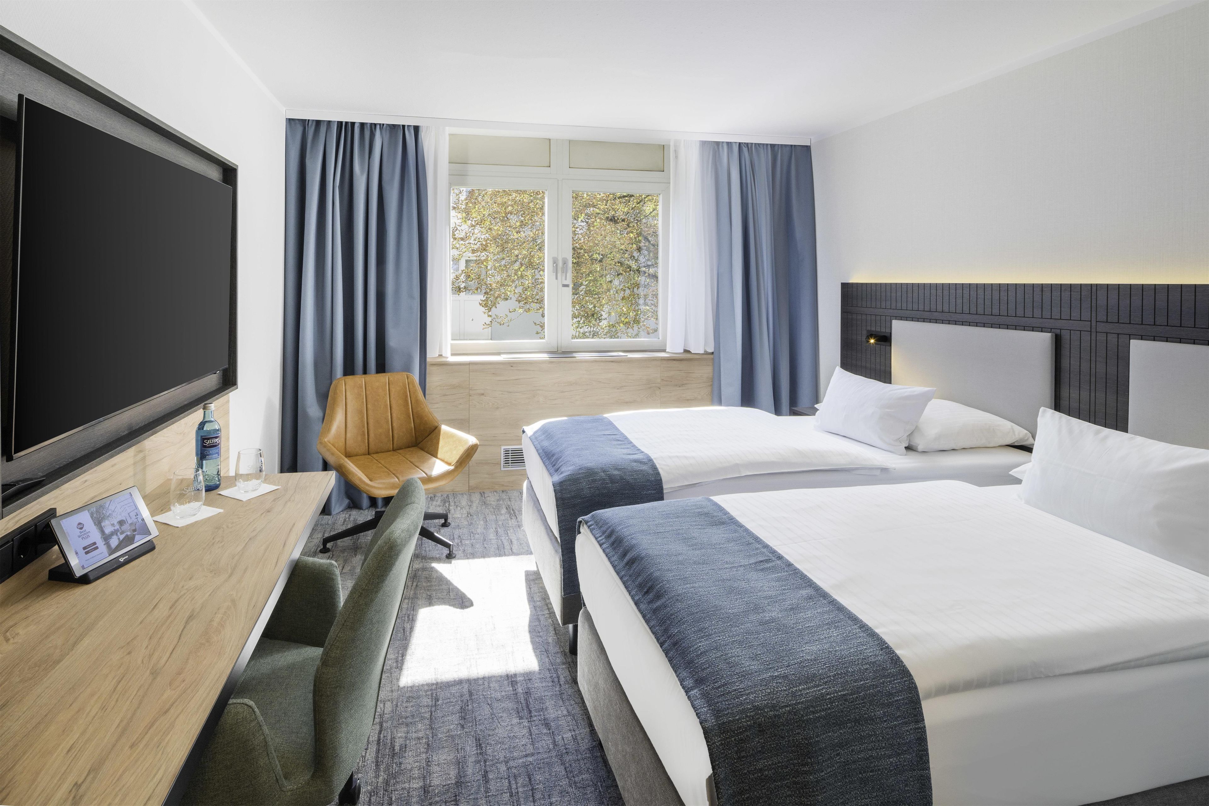 A hotel room with two beds, a desk with a TV, a chair, and a large window with curtains.