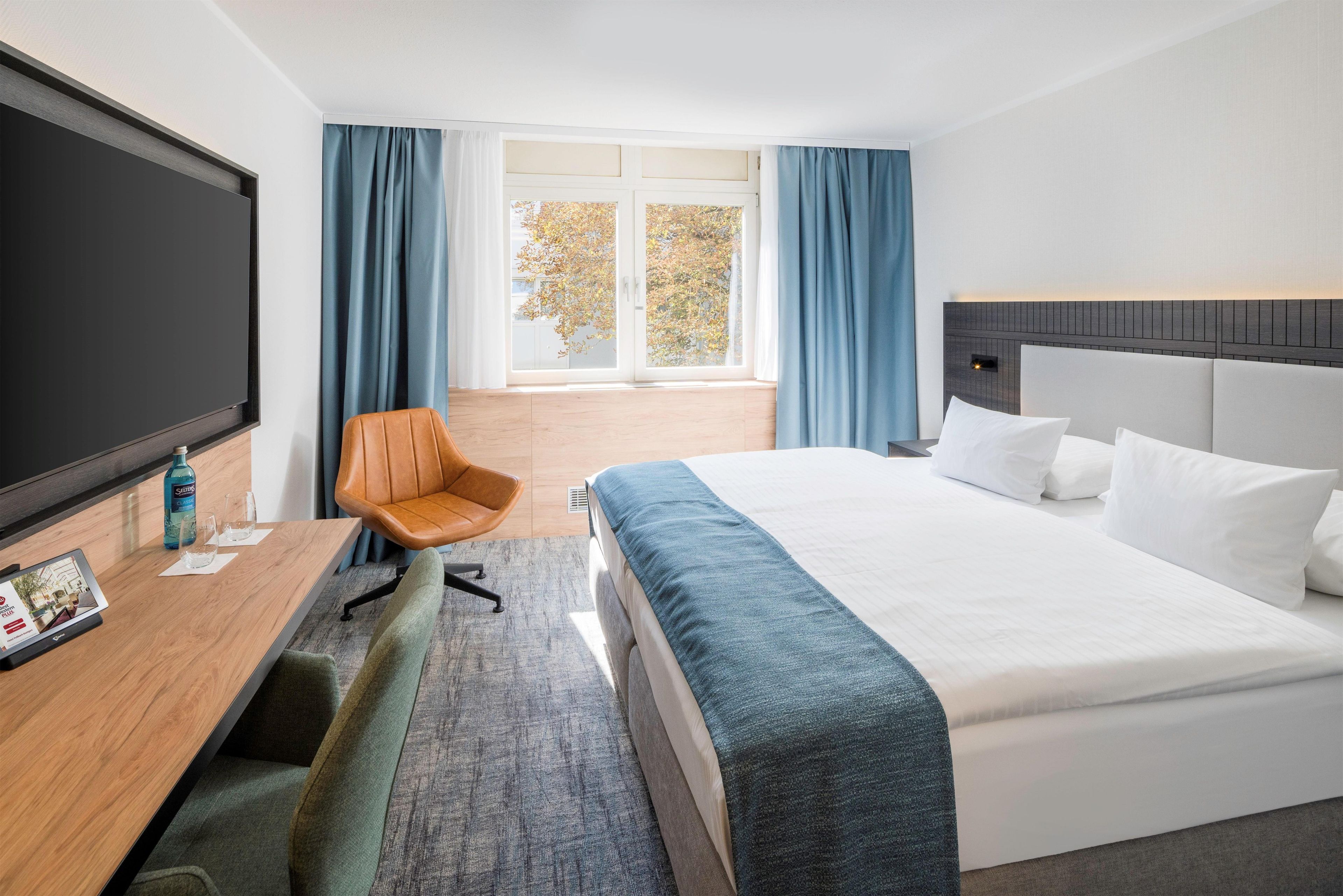 Ein Hotelzimmer mit einem großen Bett, einem Schreibtisch mit Stuhl, einem Fernseher und Blick ins Freie durch ein großes Fenster.