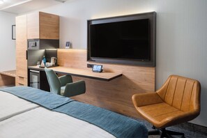 Habitación de hotel con cama, escritorio con televisor de pantalla plana, silla y una pequeña cocina.