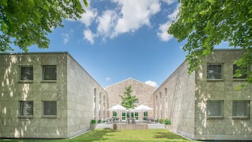 Ein Innenhof mit einem zentralen Gebäude, umgeben von zwei Flügeln mit Fenstern und einem überdachten Außenbereich zum Verweilen.