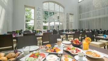 Um buffet de café da manhã com diversas opções de alimentos, incluindo frutas, pães e bebidas, situado em uma ampla área de refeições com janelas grandes e vista para o exterior.