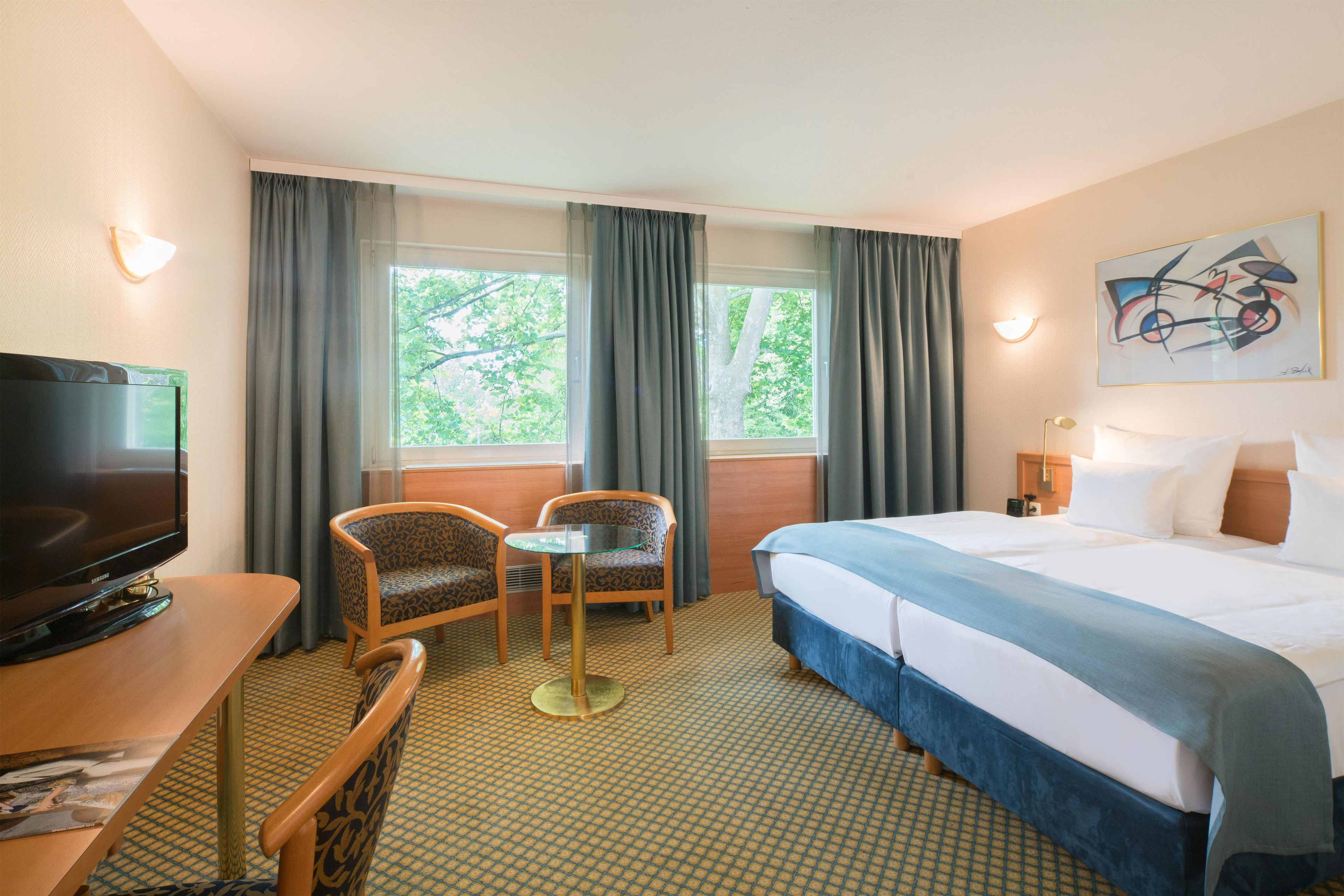 A hotel room with a bed, a desk with a TV, two chairs, a small table, and a window with curtains.