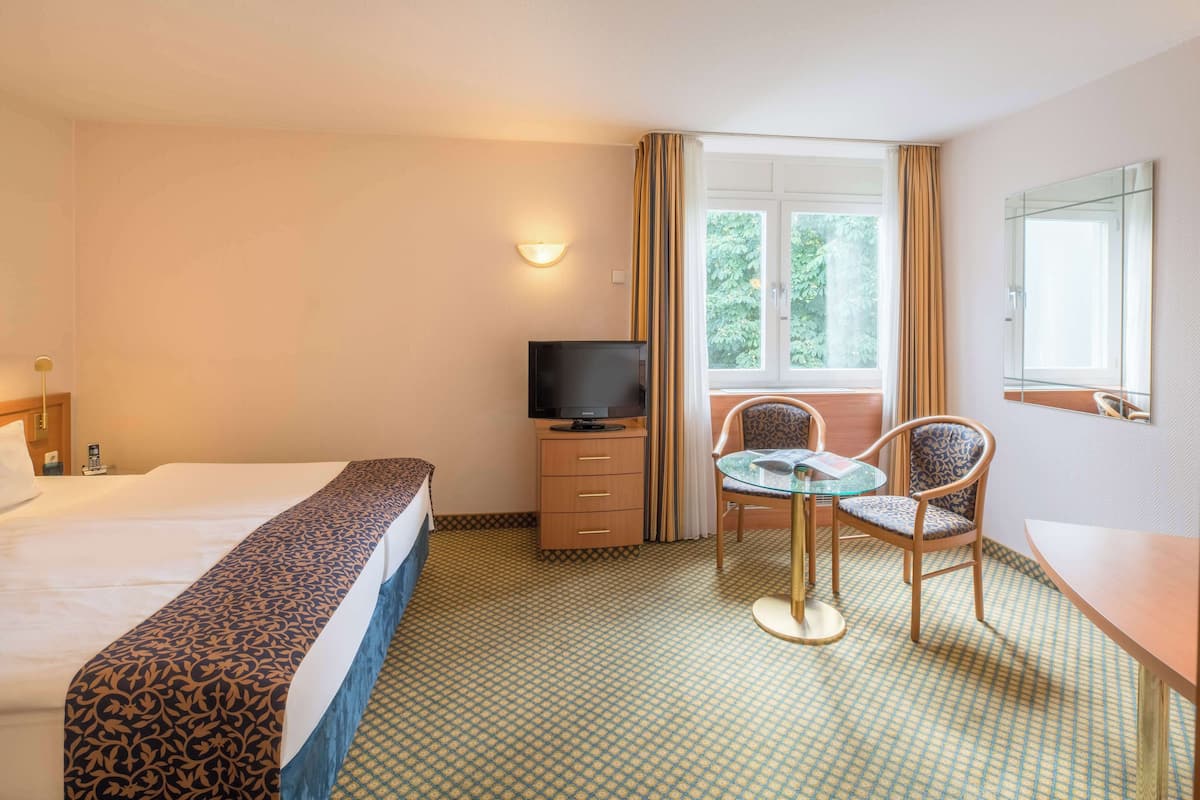 A hotel room with a bed, a TV, a desk with two chairs, and a window with curtains.