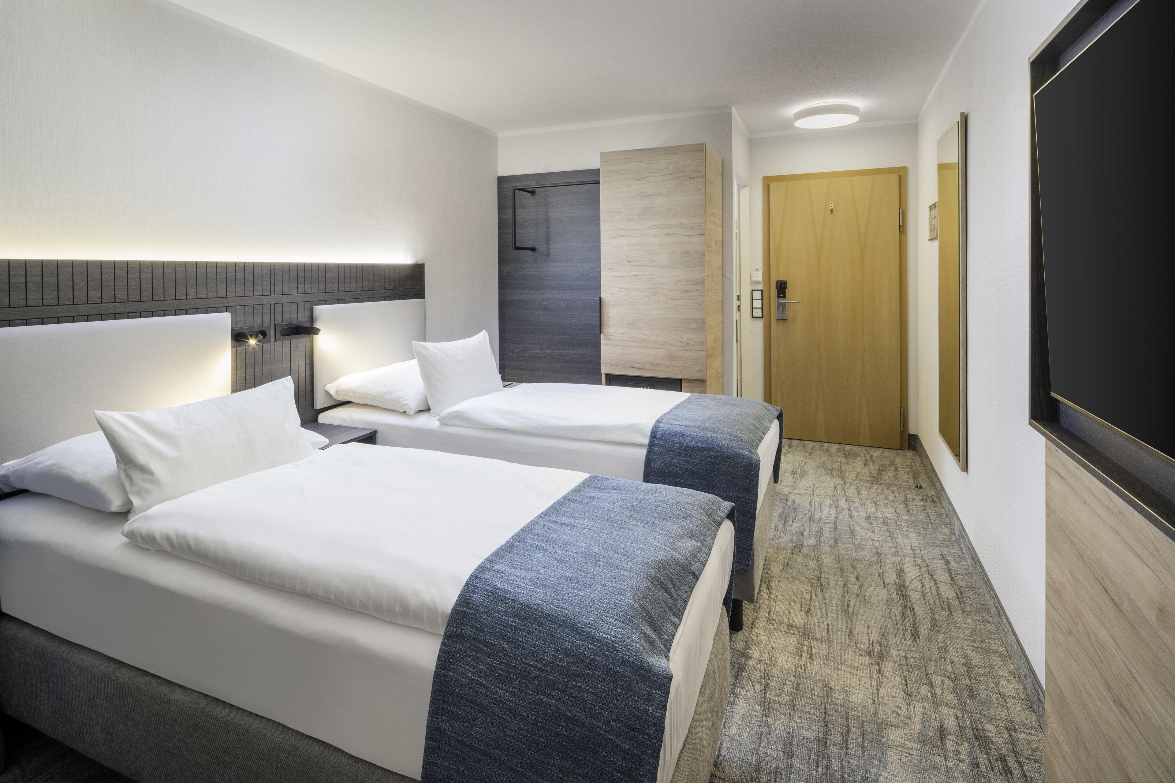 Ett hotellrum med två sängar, en trädörr, en platt-TV och ett golv täckt med matta.