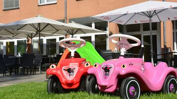 Dos coches de juguete para niños aparcados sobre césped frente a un edificio con terraza y sombrillas.