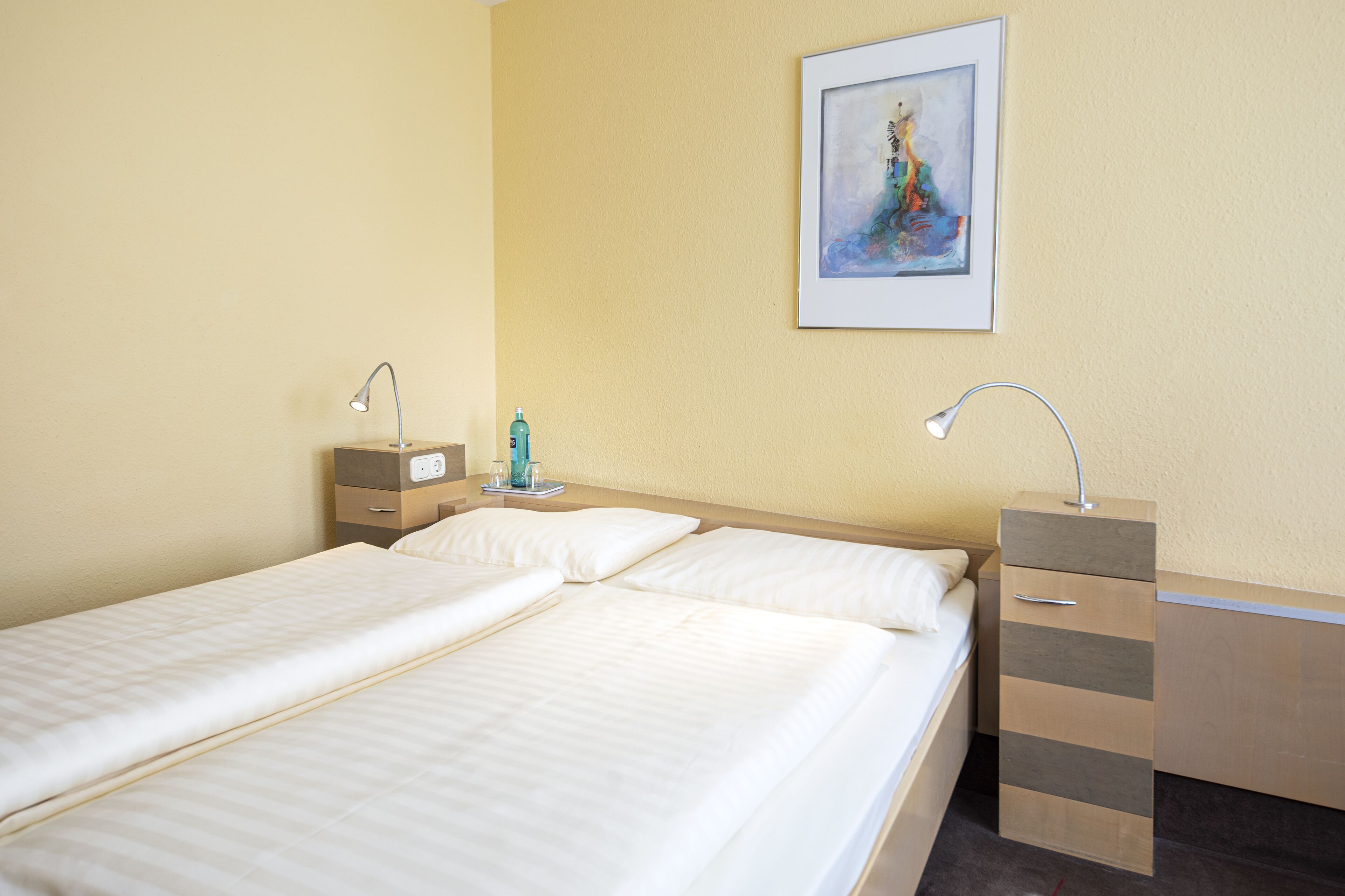 Habitación de hotel con dos camas individuales, una mesita de noche con lámpara, un archivador pequeño y un cuadro enmarcado en la pared.