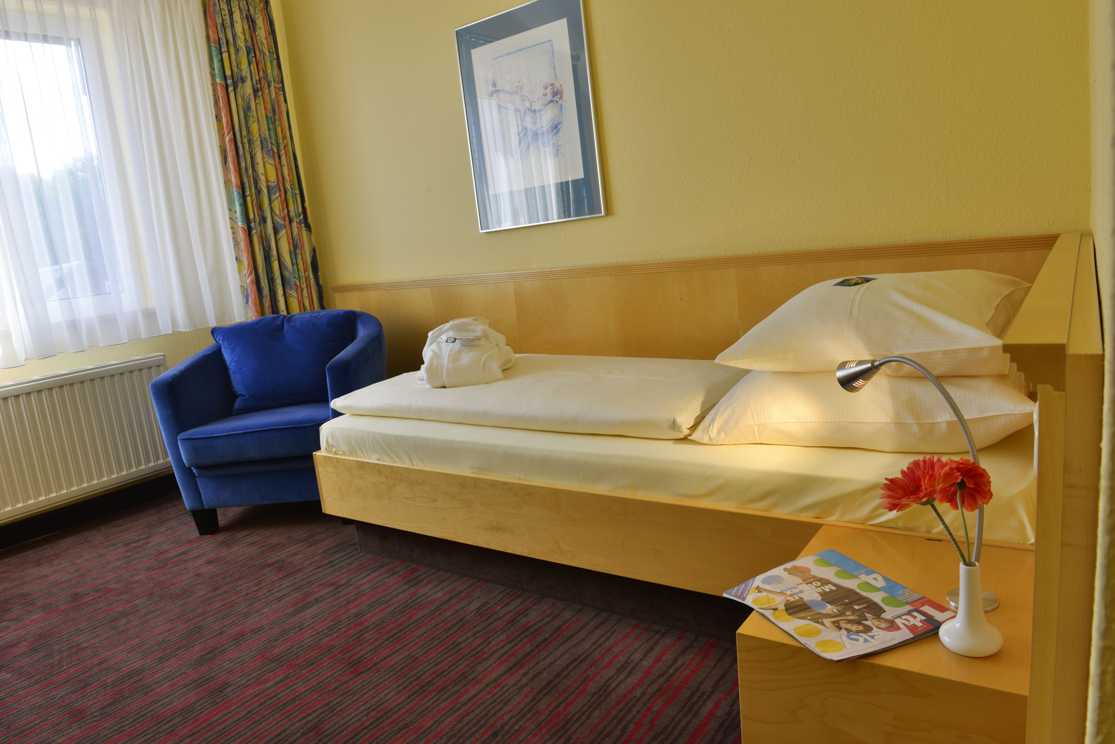 Habitación de hotel con una cama individual, un sillón azul, un radiador, una ventana con cortinas y un cuadro en la pared.