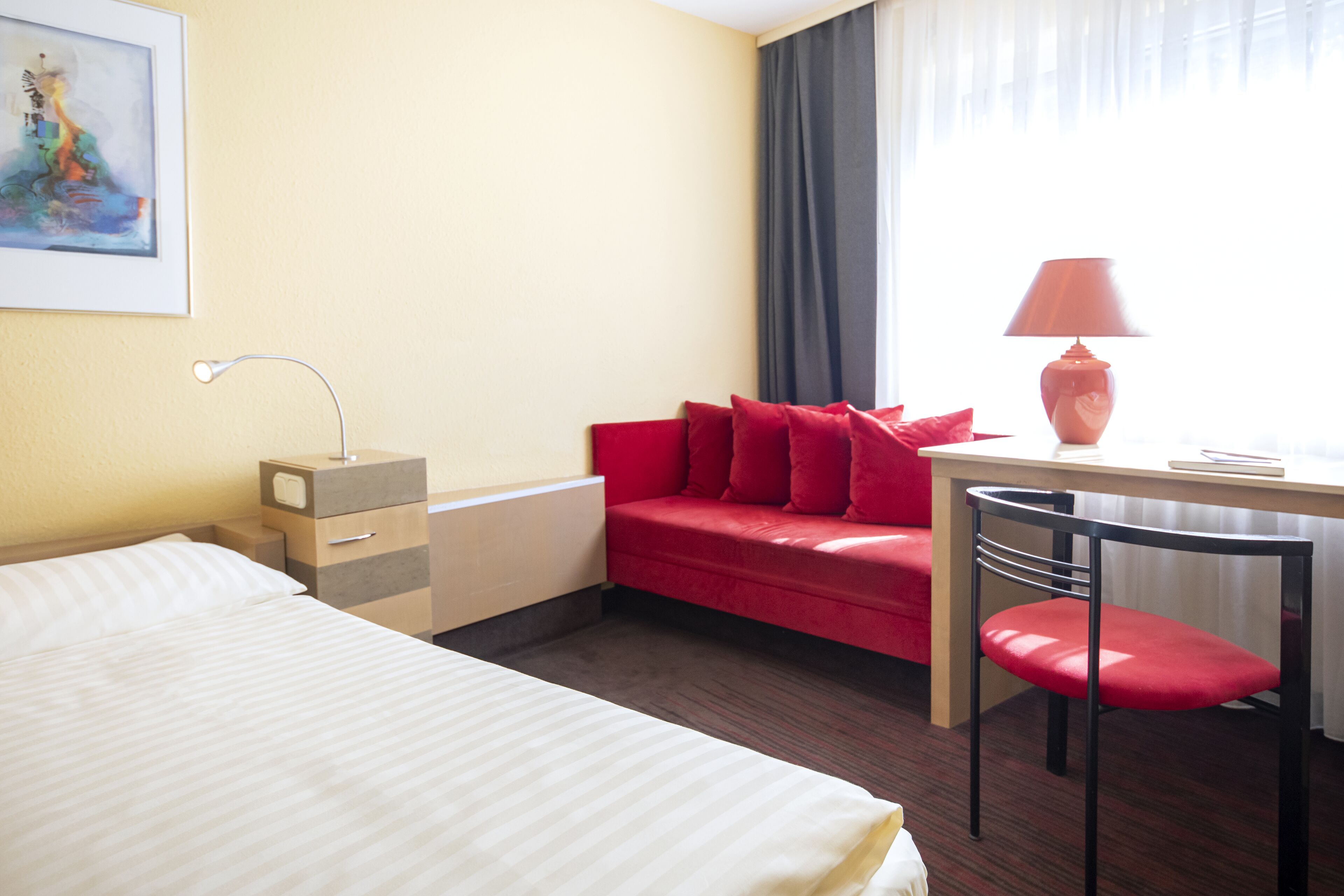 Una habitación de hotel con una cama, un sofá rojo, un escritorio con lámpara y una silla.
