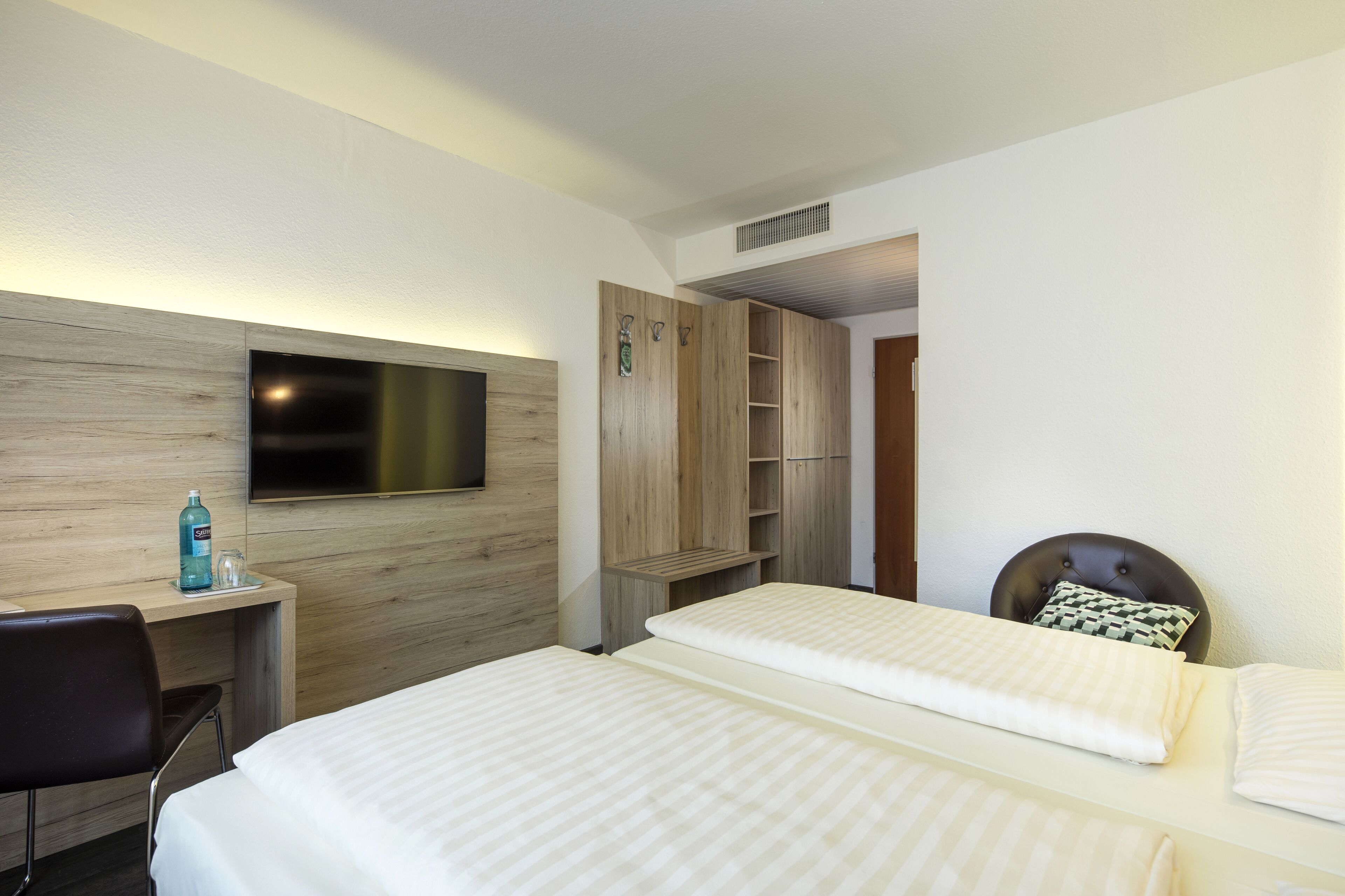 A hotel room with a bed, a desk, a chair, a TV mounted on the wall, and a wardrobe.
