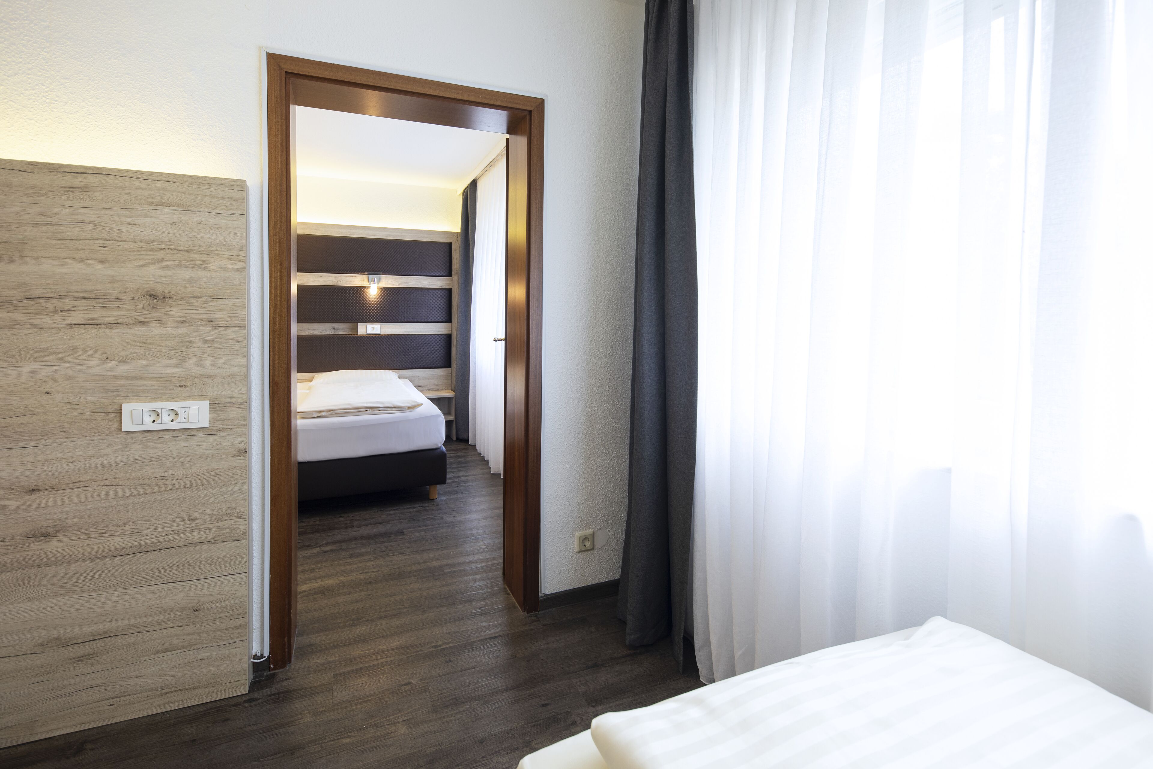 Una habitación de hotel con piso de madera, una cama con ropa blanca, una mesita de noche y un buró.