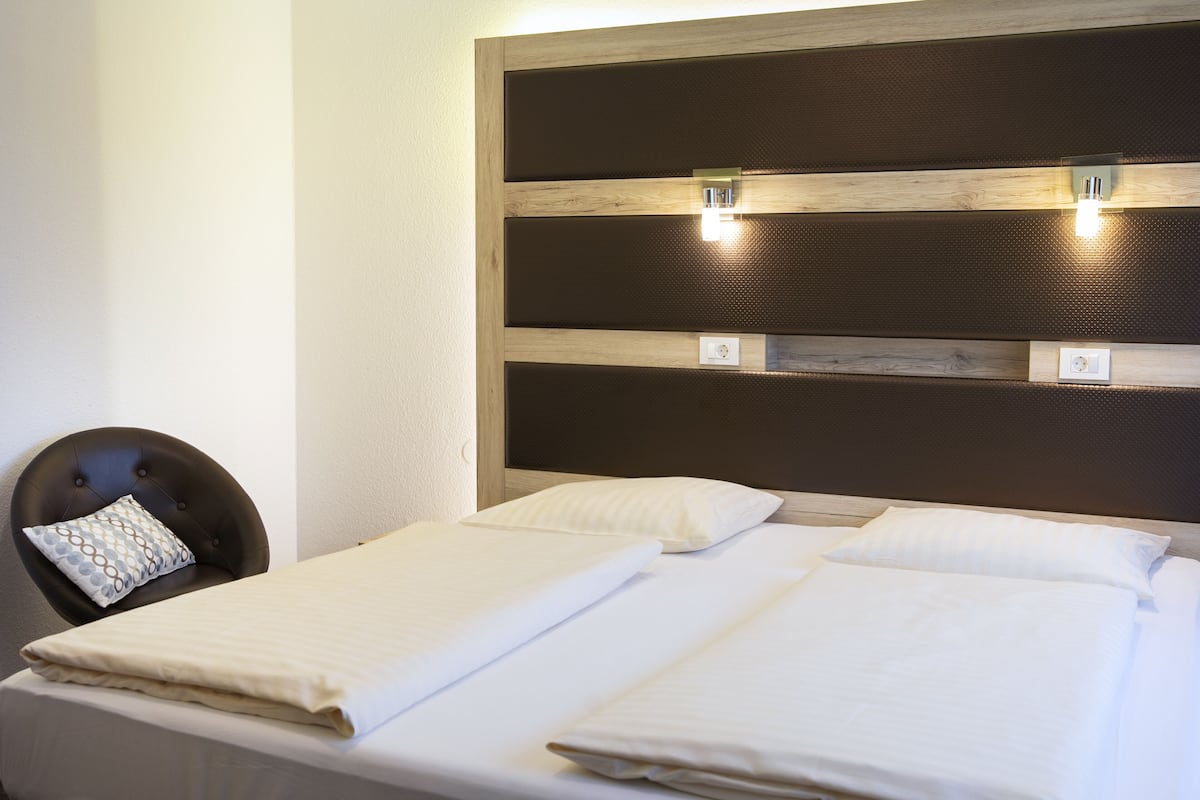 A hotel room with two beds, a headboard with built-in lighting, a chair with a patterned pillow, and a wall-mounted light fixture.