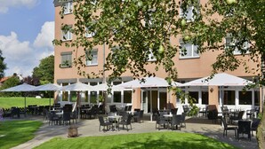 An outdoor seating area with tables and chairs, a building in the background, and a tree in the foreground. - Hotel am Park Lüneburger Heide (Hodenhagen)