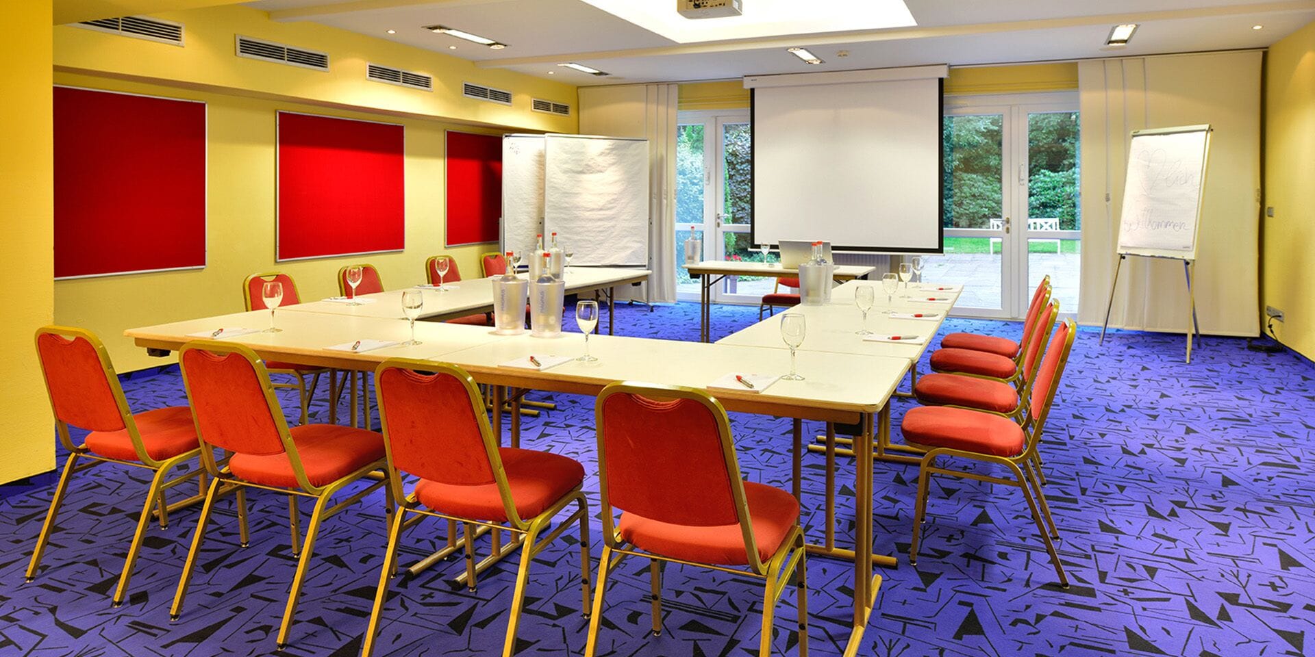 A conference room with a long table, red chairs, and a projector screen.