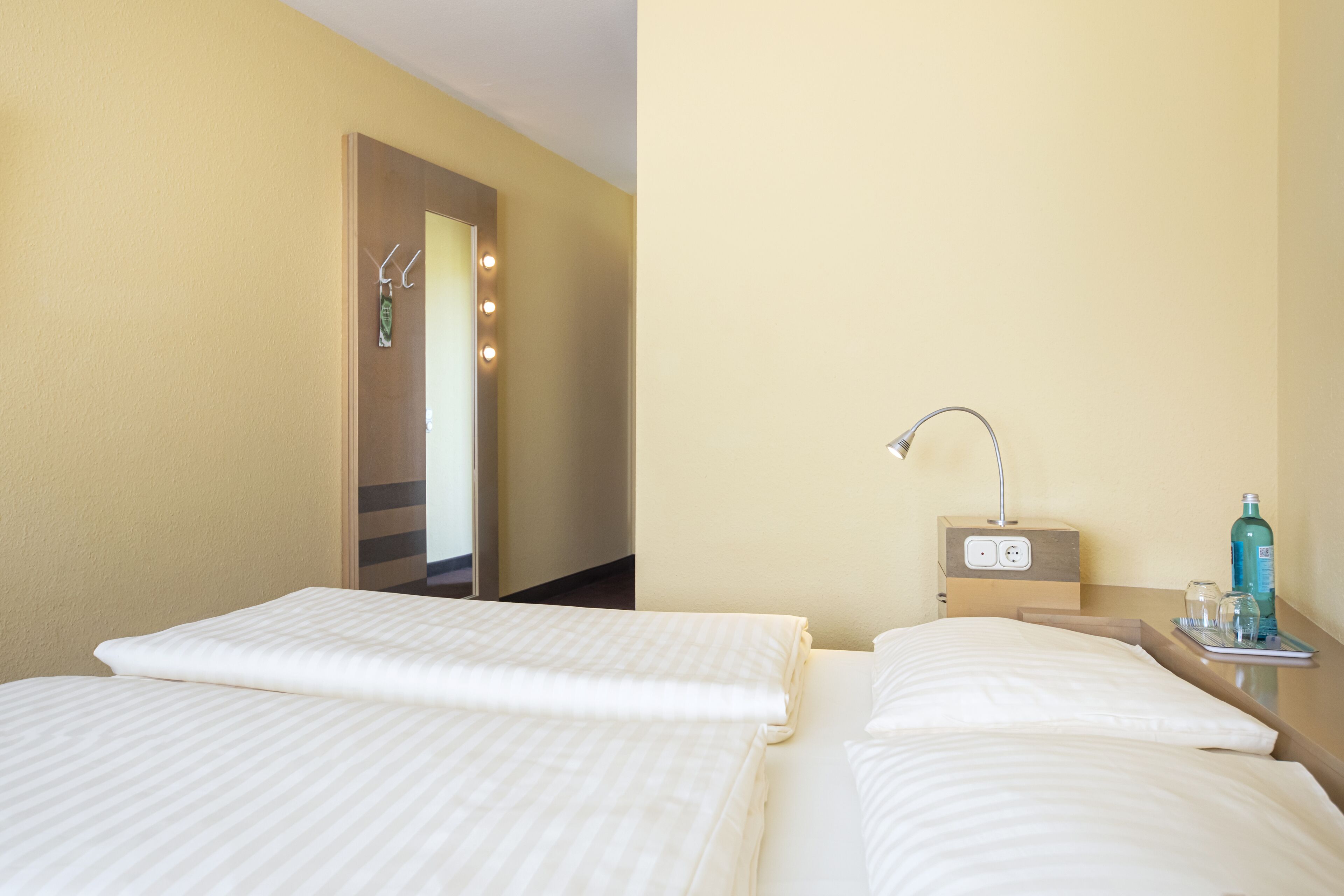 A hotel room with two beds, a bedside table with a lamp, a mirror, and a small table with a bottle and glasses.