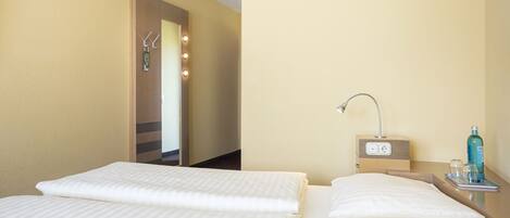 A hotel room with two beds, a bedside table with a lamp, a mirror, and a small table with a bottle and glasses.