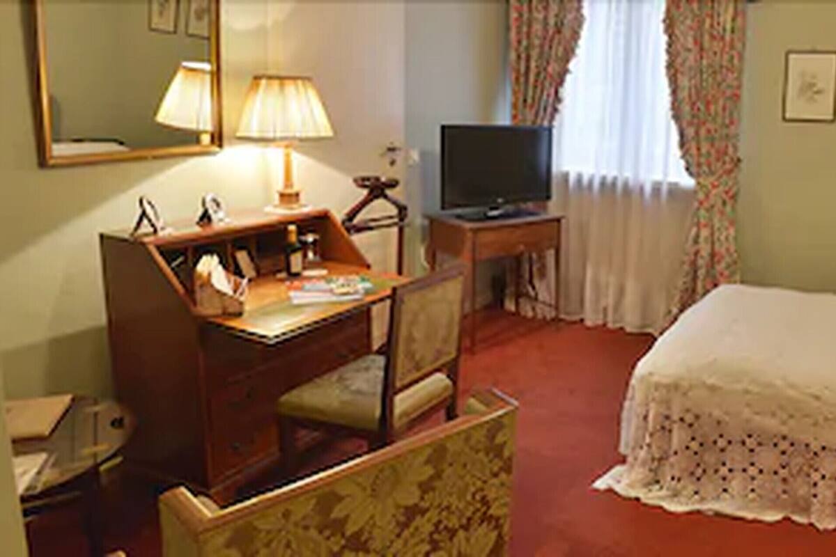 Hotellihuone, jossa on sänky, kirjoituspöytä lampun kanssa, televisio ja seinällä kehystettyjä kuvia.