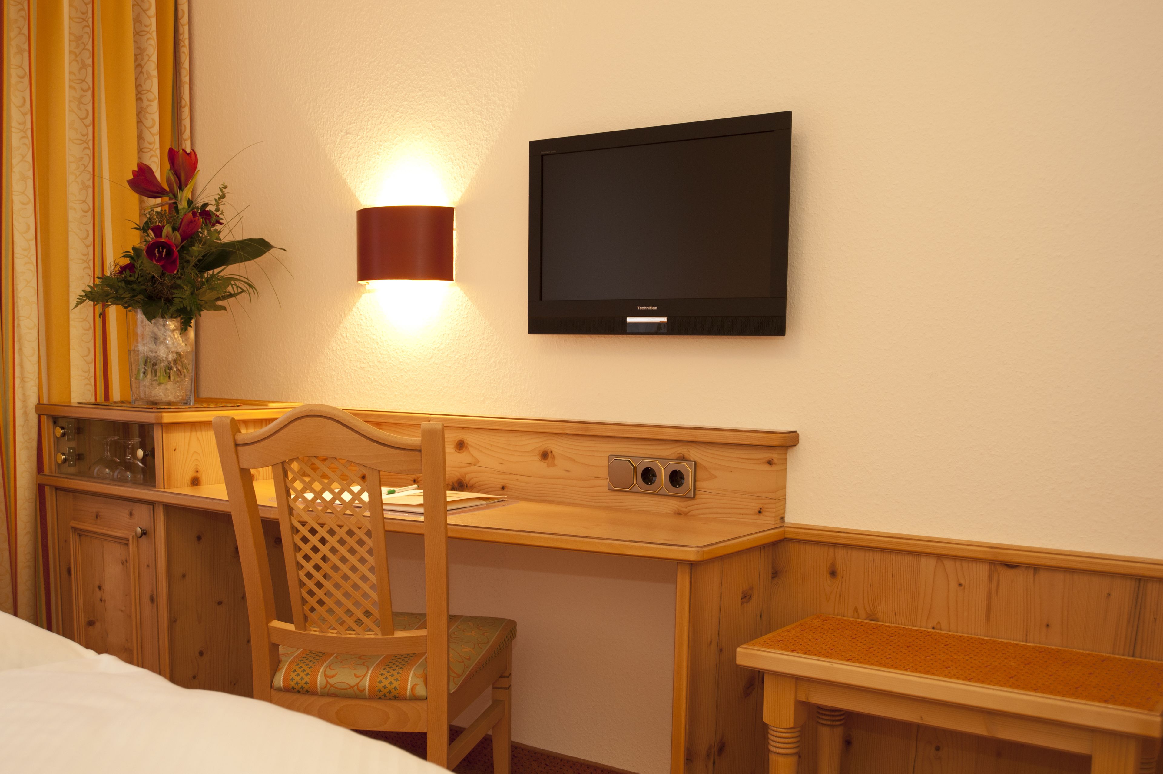 A hotel room with a wooden desk, a television mounted on the wall, a wooden chair, and a small bedside table.