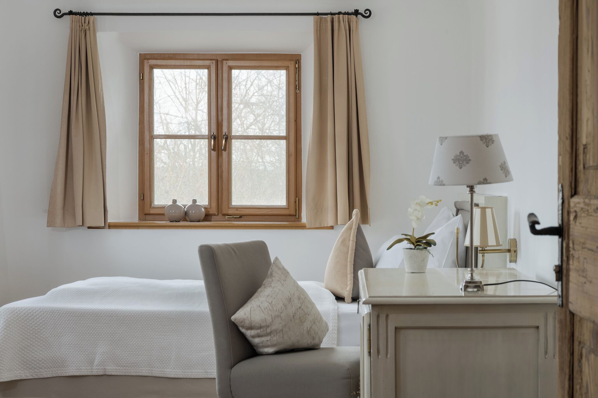 Un dormitorio con una cama, una silla, un escritorio con lámpara y una ventana con cortinas.